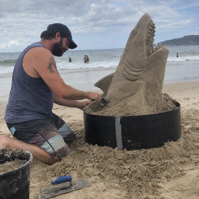 Sand sculptor using an HDPE plastic form ring to carve a shark sand sculpture on the beach
