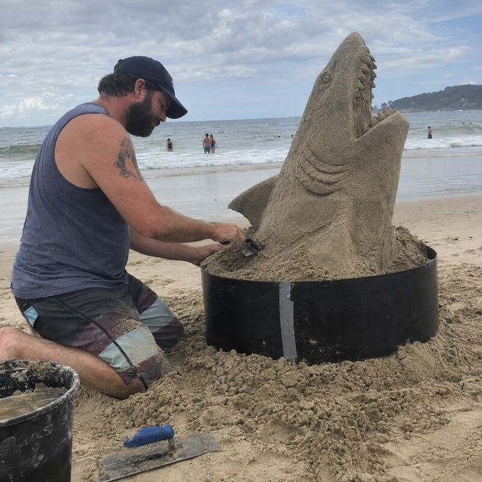 Sand sculptor using an HDPE plastic form ring to carve a shark sand sculpture on the beach
